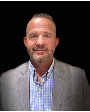 A photo of a man in a grey suit and blue checkered shirt.